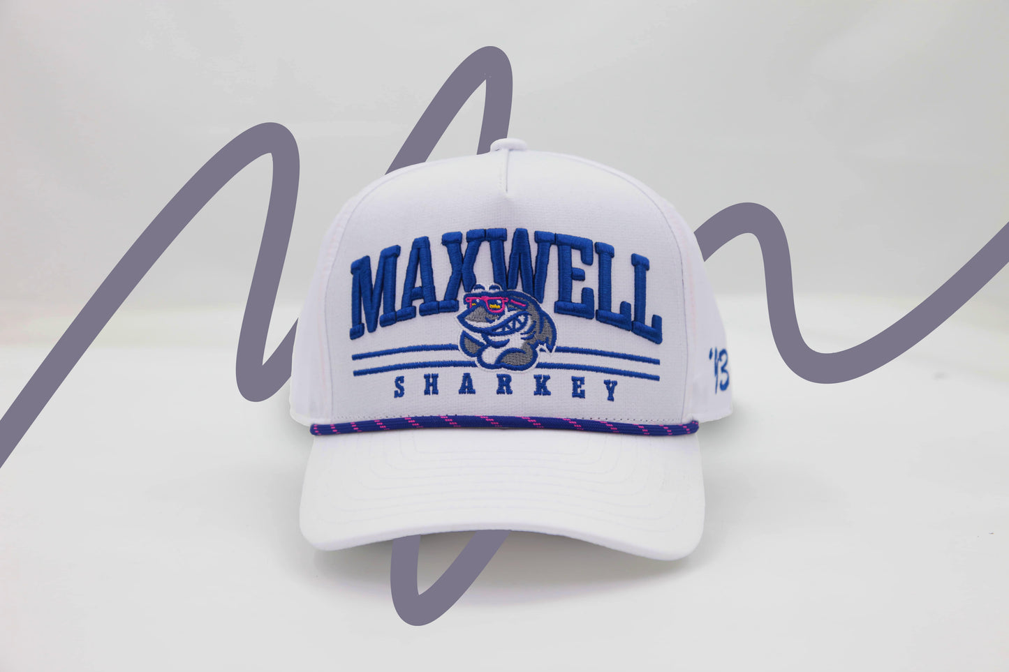 Front view of The Shark Attack Rope Hat – a bright white baseball cap embroidered with the Maxwell Sharkey mascot logo amidst bold, slab-serif letters reading "Maxwell Sharkey". It features a blue and pink rope detail across the brim.