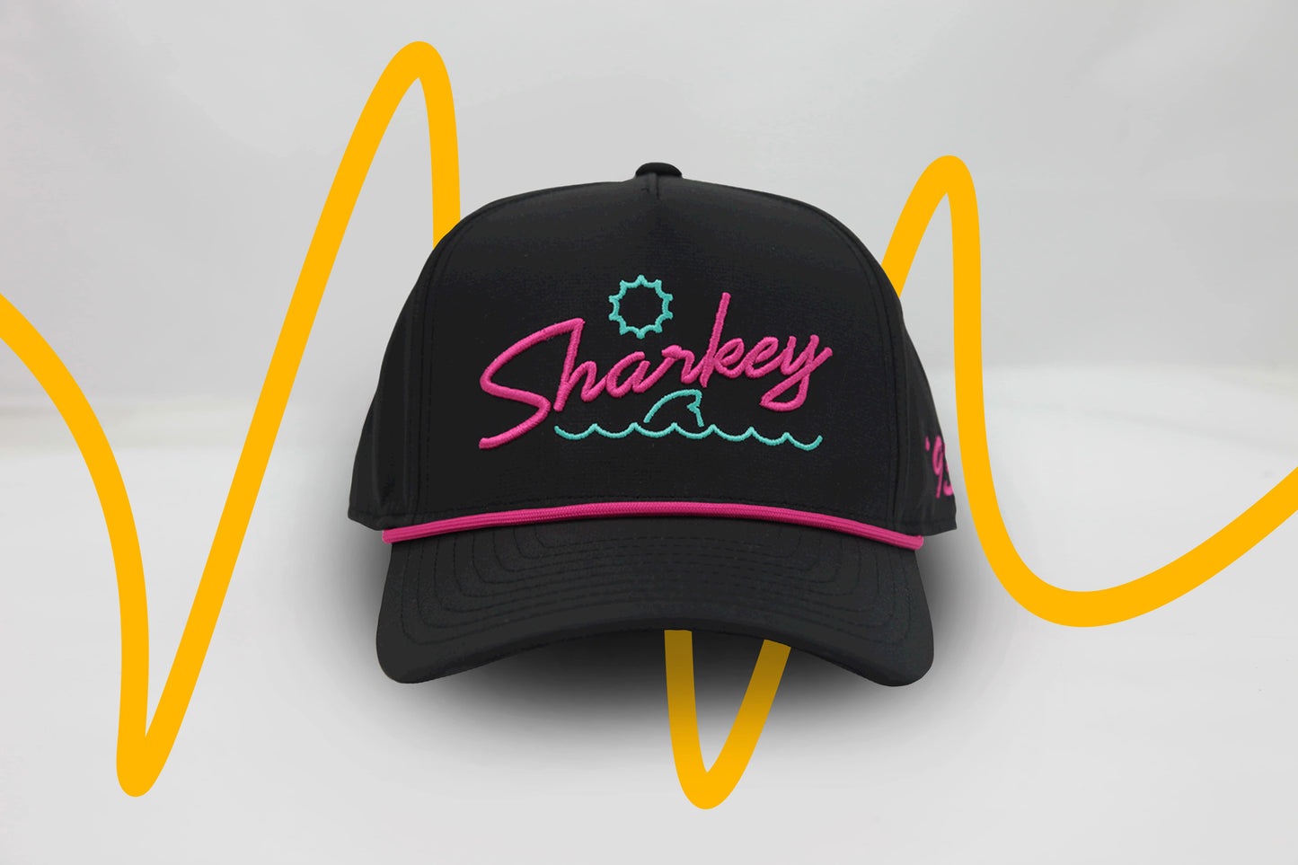 Front view of The Neon Lights Hat – black baseball cap with retro 'Sharkey' embroidery in neon pink and teal, sun and wave graphic, and pink rope detail above brim.
