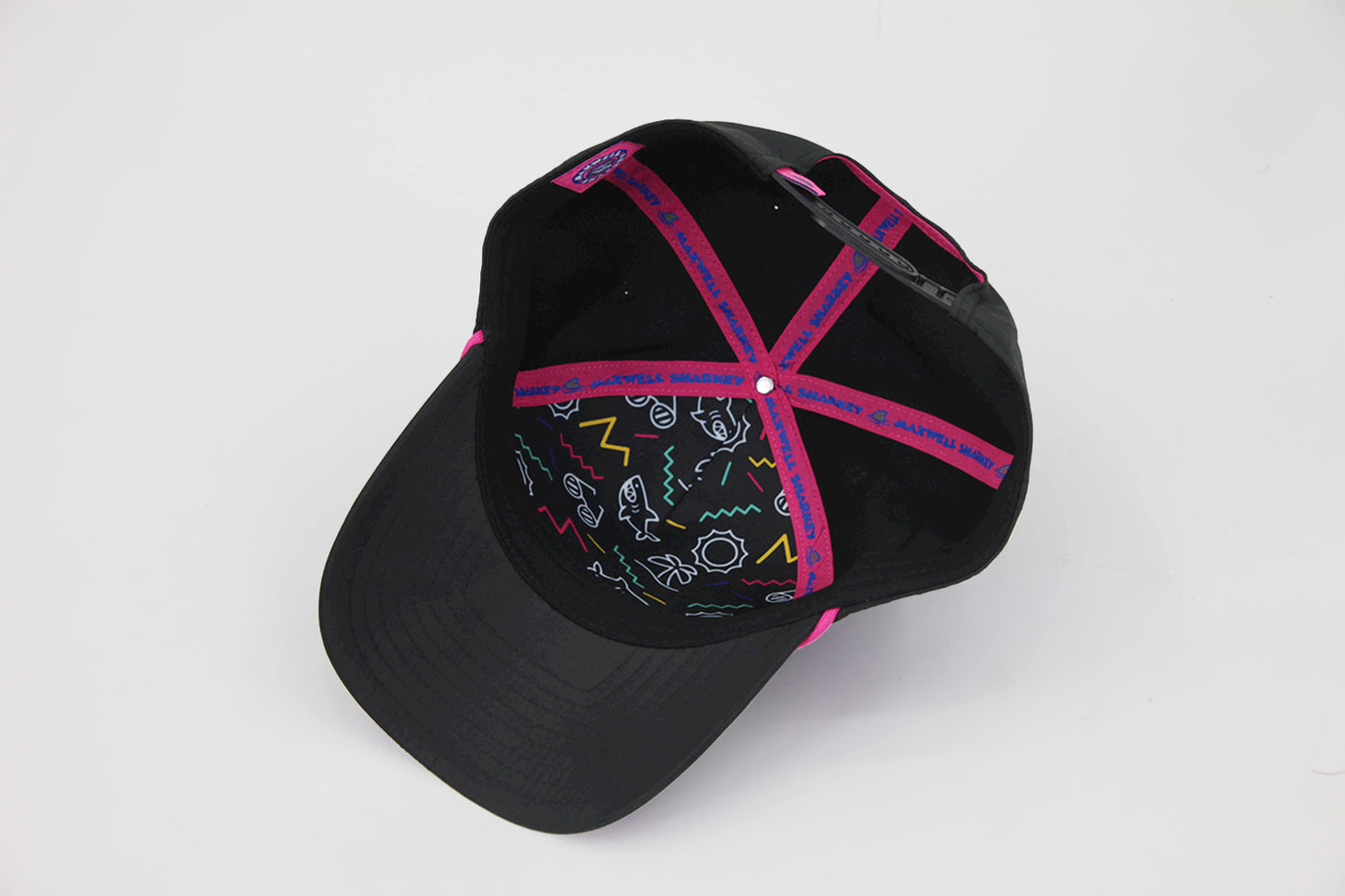 Inside view of a black baseball cap with neon pink interior stitching, Maxwell Sharkey branding on the taping, and colorful 90s-style graphic lining under the crown.