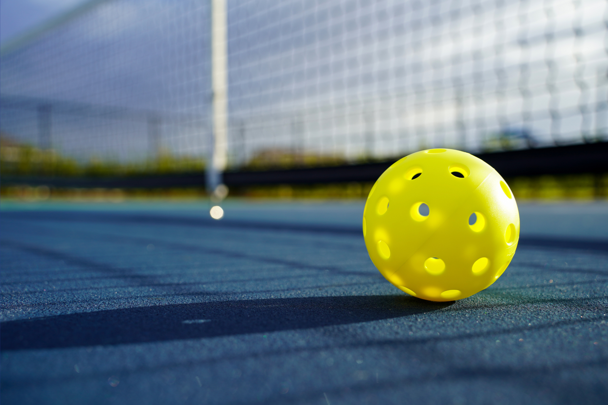 Yellow pickleball resting on a blue court near the net, ready for action under bright sunlight.