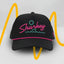 Front view of The Neon Lights Hat – black baseball cap with retro 'Sharkey' embroidery in neon pink and teal, sun and wave graphic, and pink rope detail above brim.
