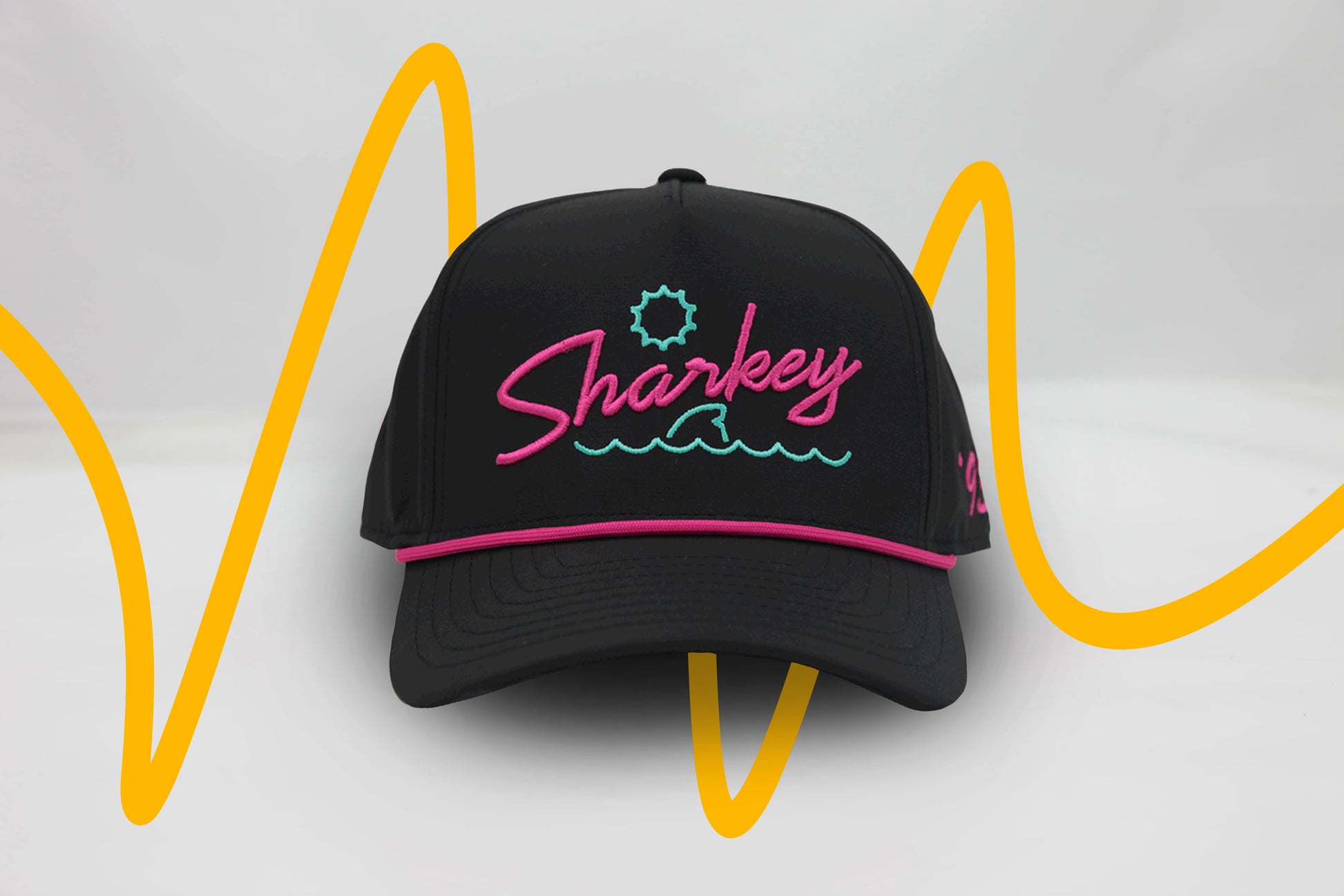Front view of The Neon Lights Hat – black baseball cap with retro 'Sharkey' embroidery in neon pink and teal, sun and wave graphic, and pink rope detail above brim.